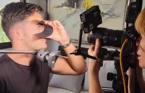 Iridology photo setup: subject holding a lens cup to his eye while a photographer aims a DSLR on a tripod